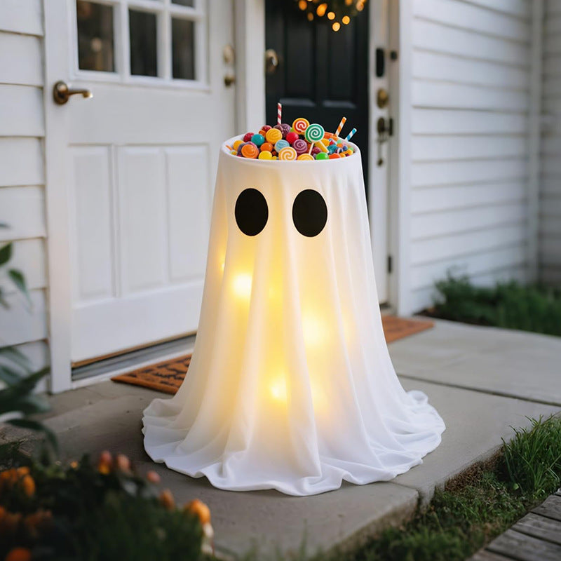 A ghost-shaped table lamp or decoration draped in white fabric stands on a front porch. The top of the ghost is filled with colorful lollipops and candies, and the body glows with warm yellow light.