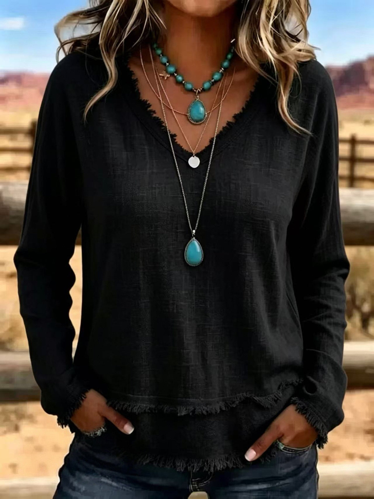 A woman wearing a black long-sleeve V-neck top with a frayed hem, layered with turquoise and silver necklaces, standing outdoors in a desert landscape.