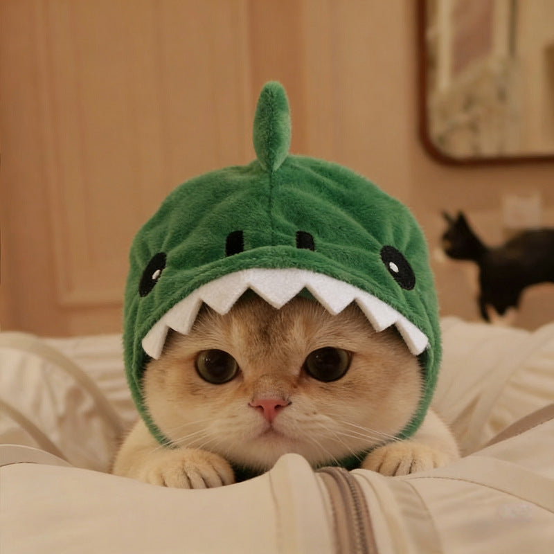 A cream-colored cat wearing a plush green dinosaur shark hat with white teeth, resting on a white bed in a warm indoor setting.