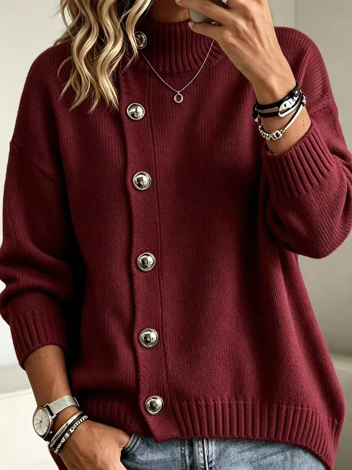 A woman wearing a burgundy knit cardigan sweater with large silver buttons, paired with light blue jeans. She is shown in a close-up selfie-style shot, wearing jewelry and bracelets.