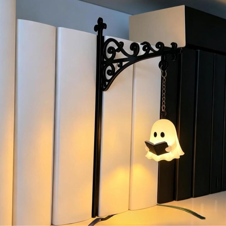 A decorative black metal wall bracket holding a hanging ghost-shaped night light that reads a book, shown against a background of large white and black books.