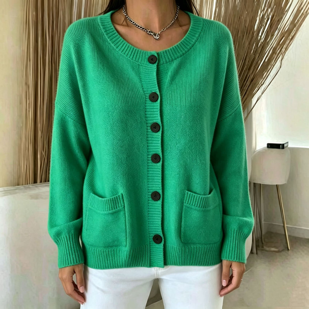 A woman wearing a bright green ribbed knit cardigan sweater with dark buttons and front pockets, paired with white pants, standing indoors.