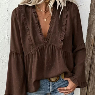 A woman wearing a dark brown long-sleeve blouse with ruffle trim and a deep V-neck, paired with blue jeans and gold necklaces, standing against a white textured wall.