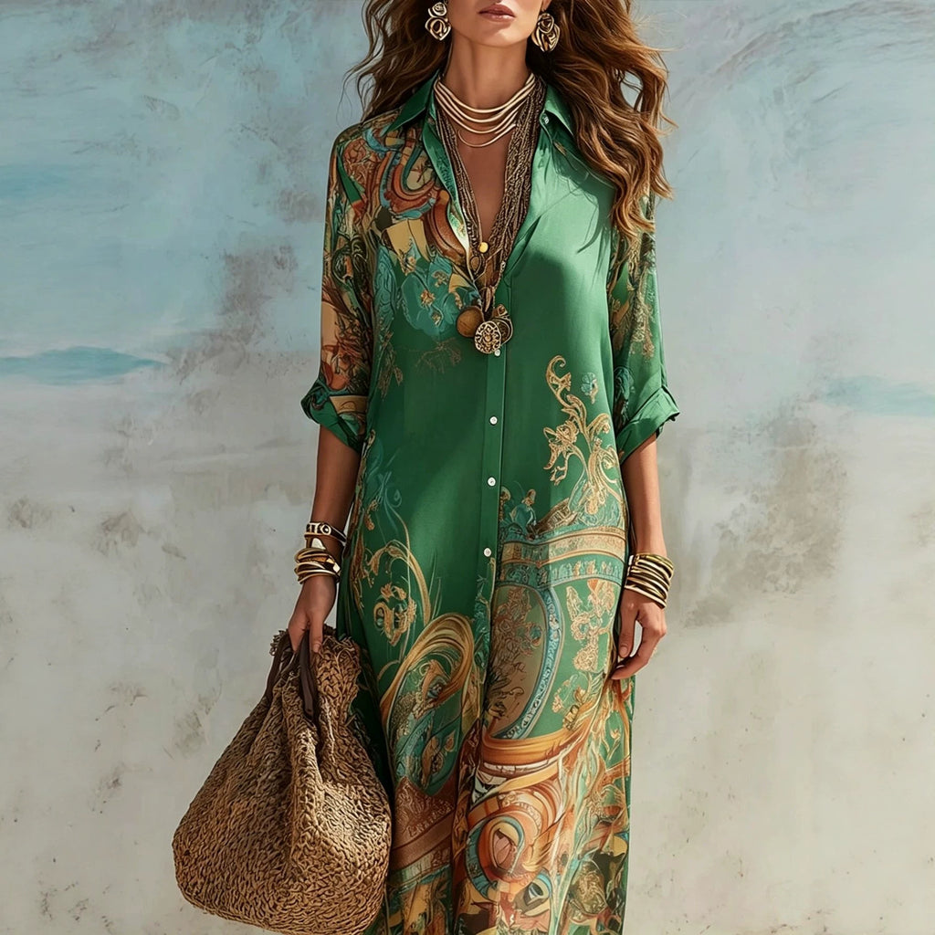 A woman models a long-sleeve button-front shirt dress with a green base and an ornate paisley-style print in gold, brown, and teal. She is accessorized with layered necklaces, bracelets, and a woven straw tote bag, standing against a textured light blue and gray wall.