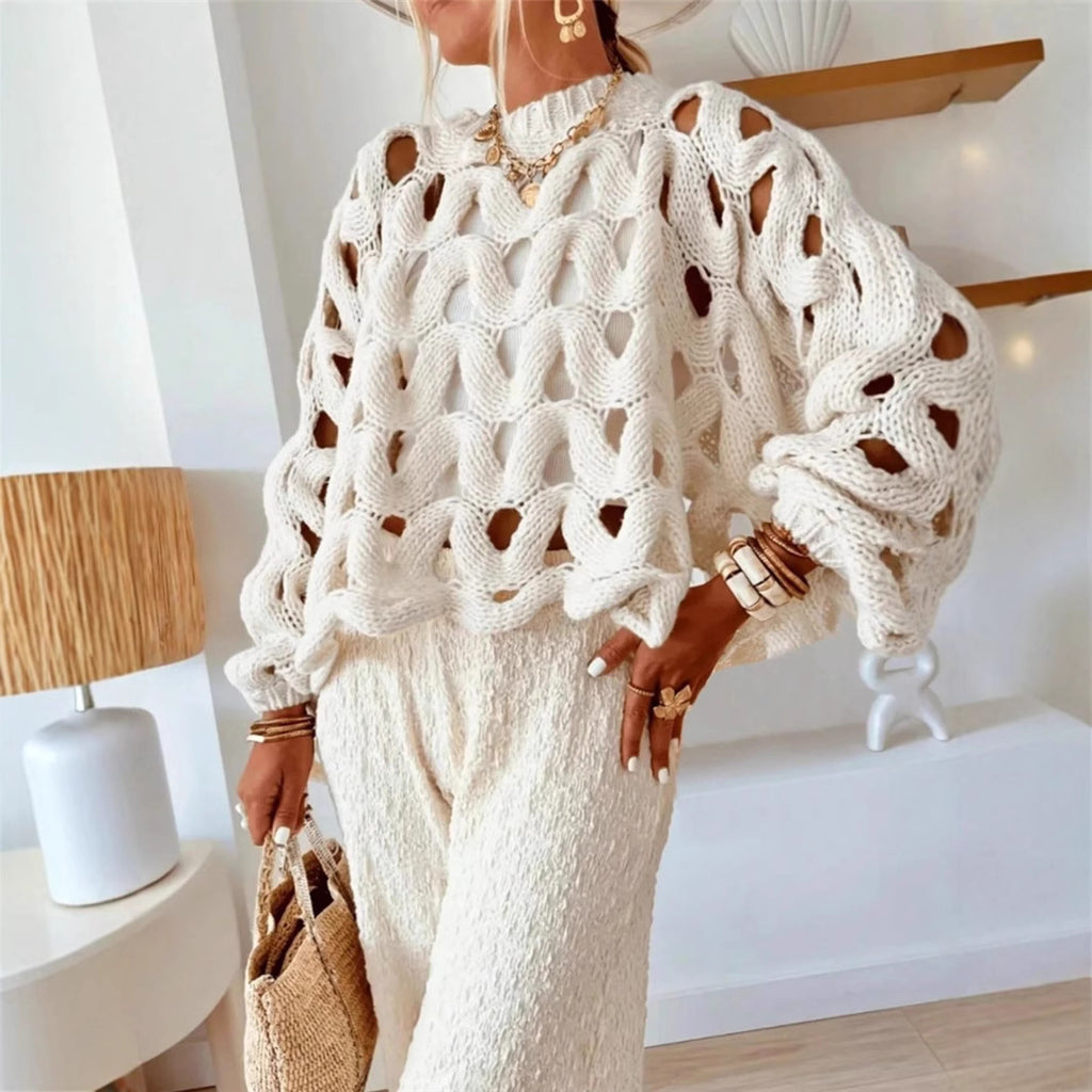 Person wearing a white crochet sweater and pants in a room with a lamp and shelf.