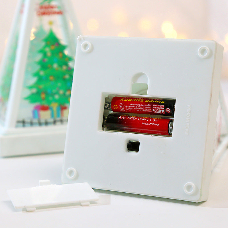 A white plastic battery-operated Christmas light box with its lid removed, showing two AAA batteries inside. A blurred festive background with a decorated Christmas tree and warm bokeh lights is visible behind it.