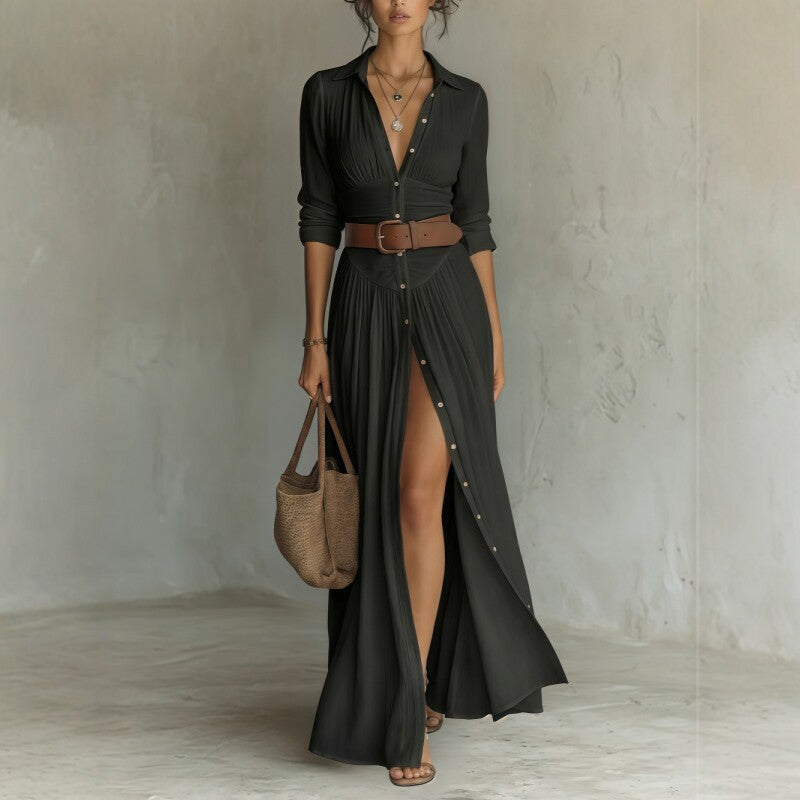 Woman wearing a long black dress with a belt, holding a woven bag against a plain background