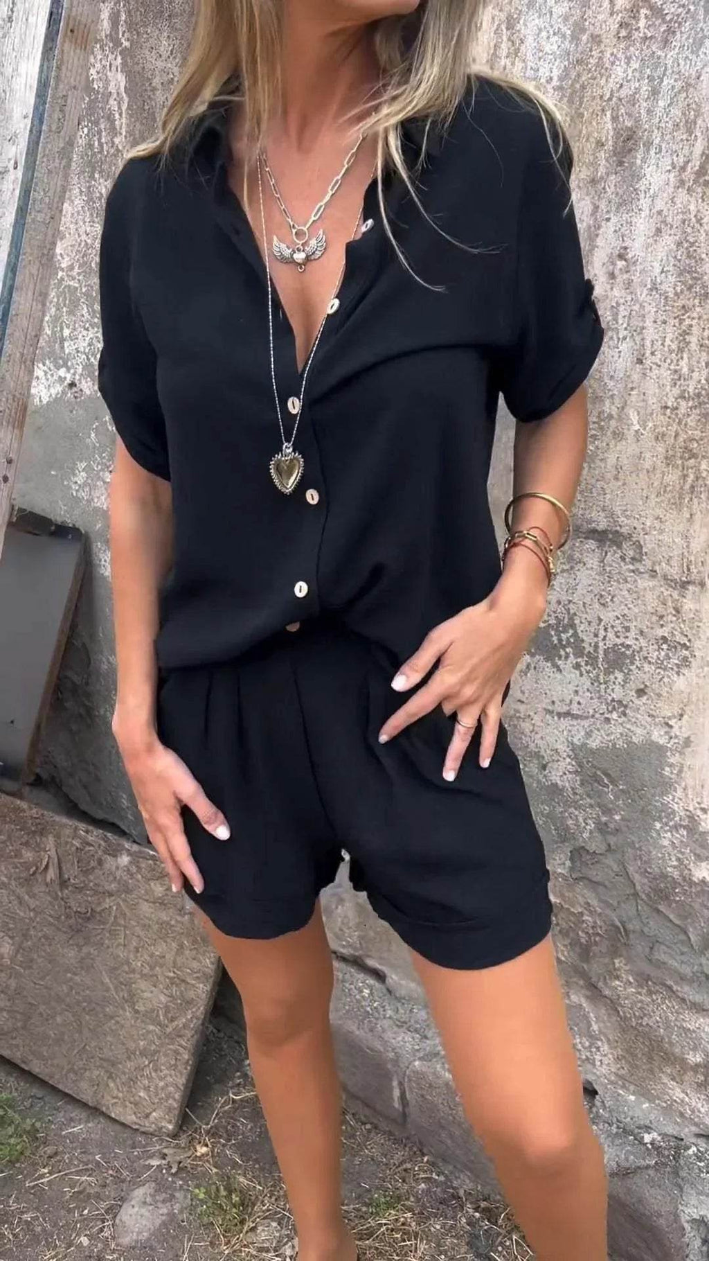Woman wearing trendy black button-up shirt and shorts set from ZA-Zola, accessorized with layered silver necklaces and bracelets, standing against a rustic wall.