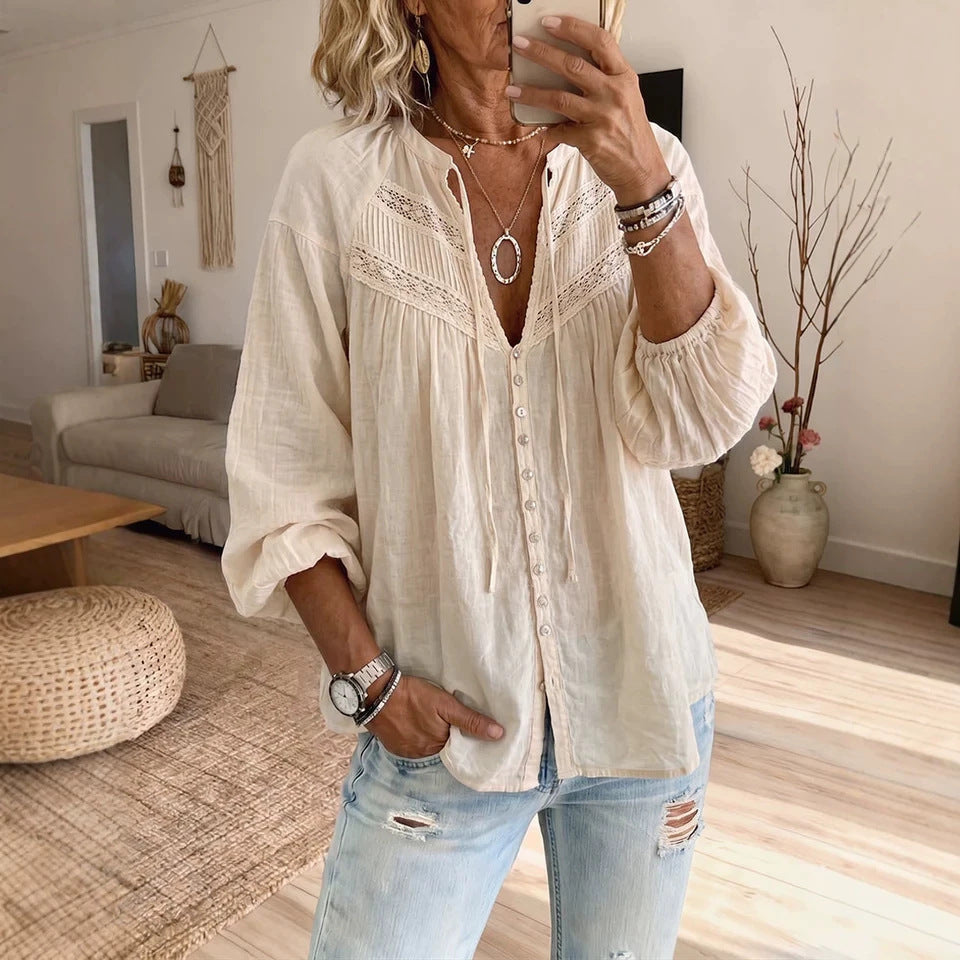 A woman takes a mirror selfie in a cozy living room, wearing a cream-colored long-sleeve button-up blouse with delicate lace detailing at the neckline and light-wash distressed jeans.