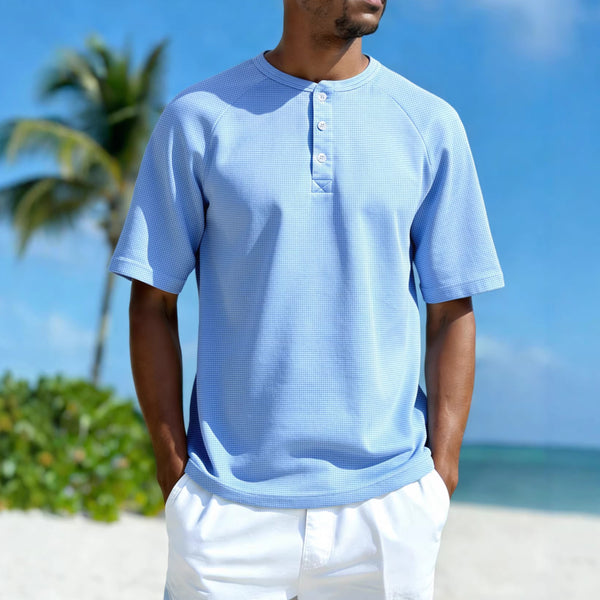 Man wearing a light blue shirt and white pants standing on a beach with palm trees and blue sky in the background.
