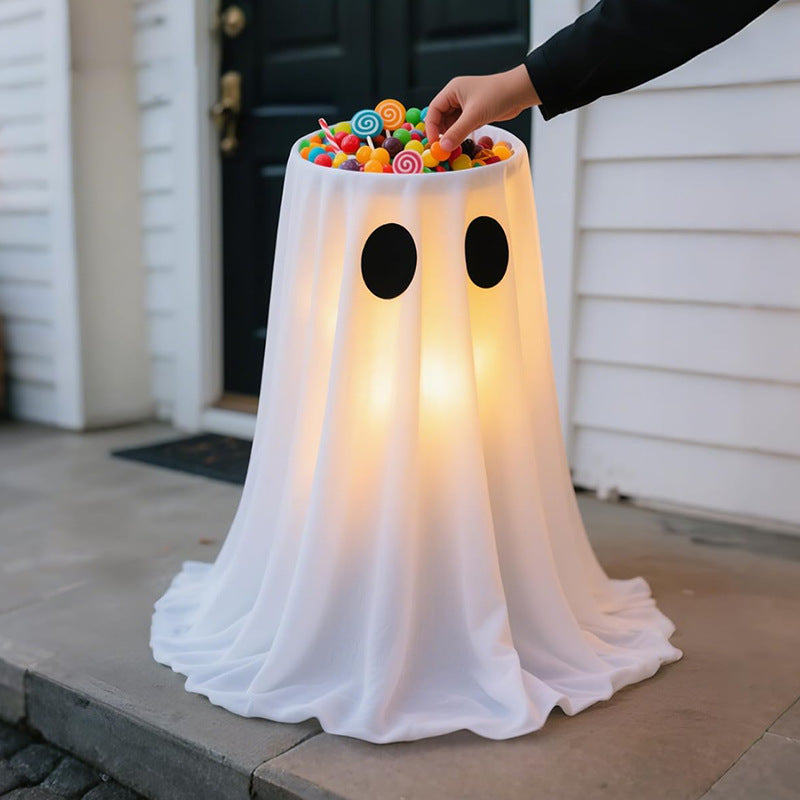 A ghost-shaped outdoor light fixture draped in white fabric, with black circular eyes and a top opening filled with colorful lollipops and candies. A person's arm is reaching in from the right to adjust the candy, set against a residential porch background.