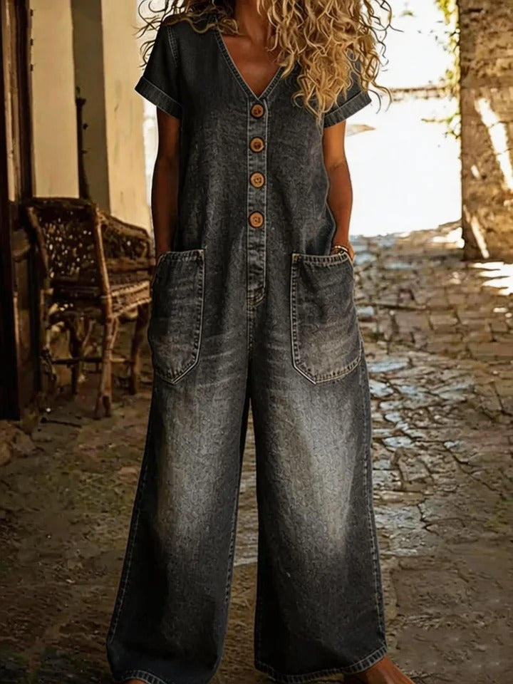 Person wearing a denim jumpsuit standing in an outdoor setting with stone walls and a bench.