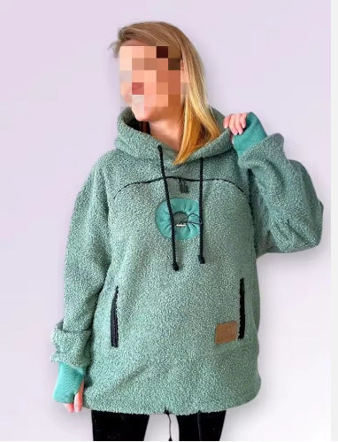 A person wearing a fuzzy sage green hooded sweatshirt with black drawstrings, black zipper details, and a small brown patch on the lower front, against a pale lavender background.
