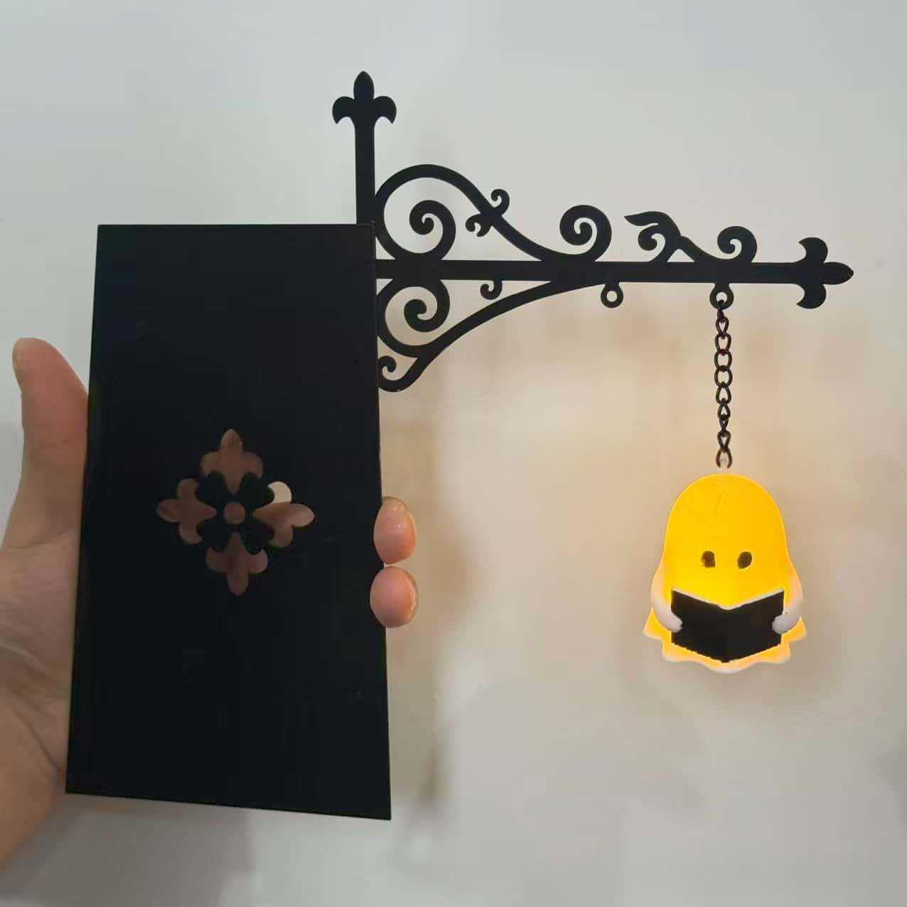 A hand holds a black decorative wall sign with an ornate scrollwork bracket and hanging chain. The sign has a rectangular black panel with a floral cutout and hangs next to a small yellow ghost-shaped light or ornament reading 'I AM A GHOST'.