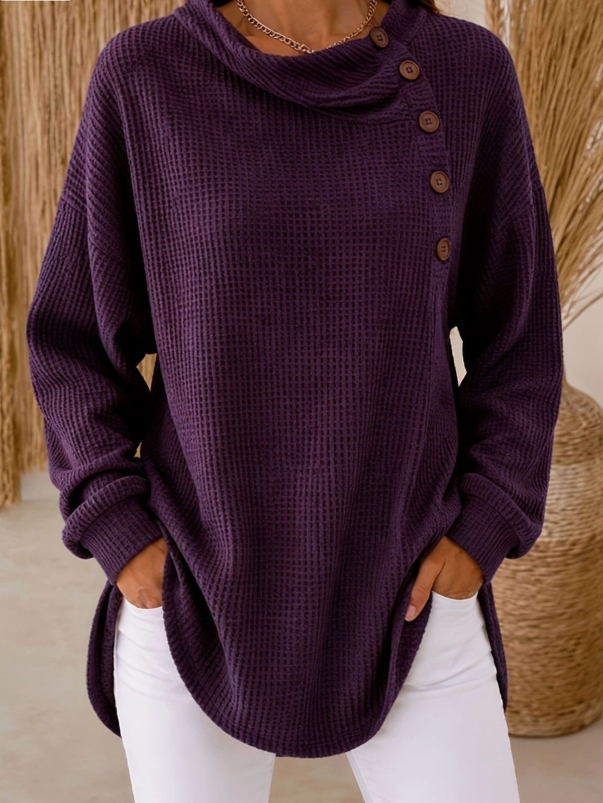 A person wearing a dark purple waffle-knit cowl-neck sweater with an asymmetrical button closure, paired with white pants.