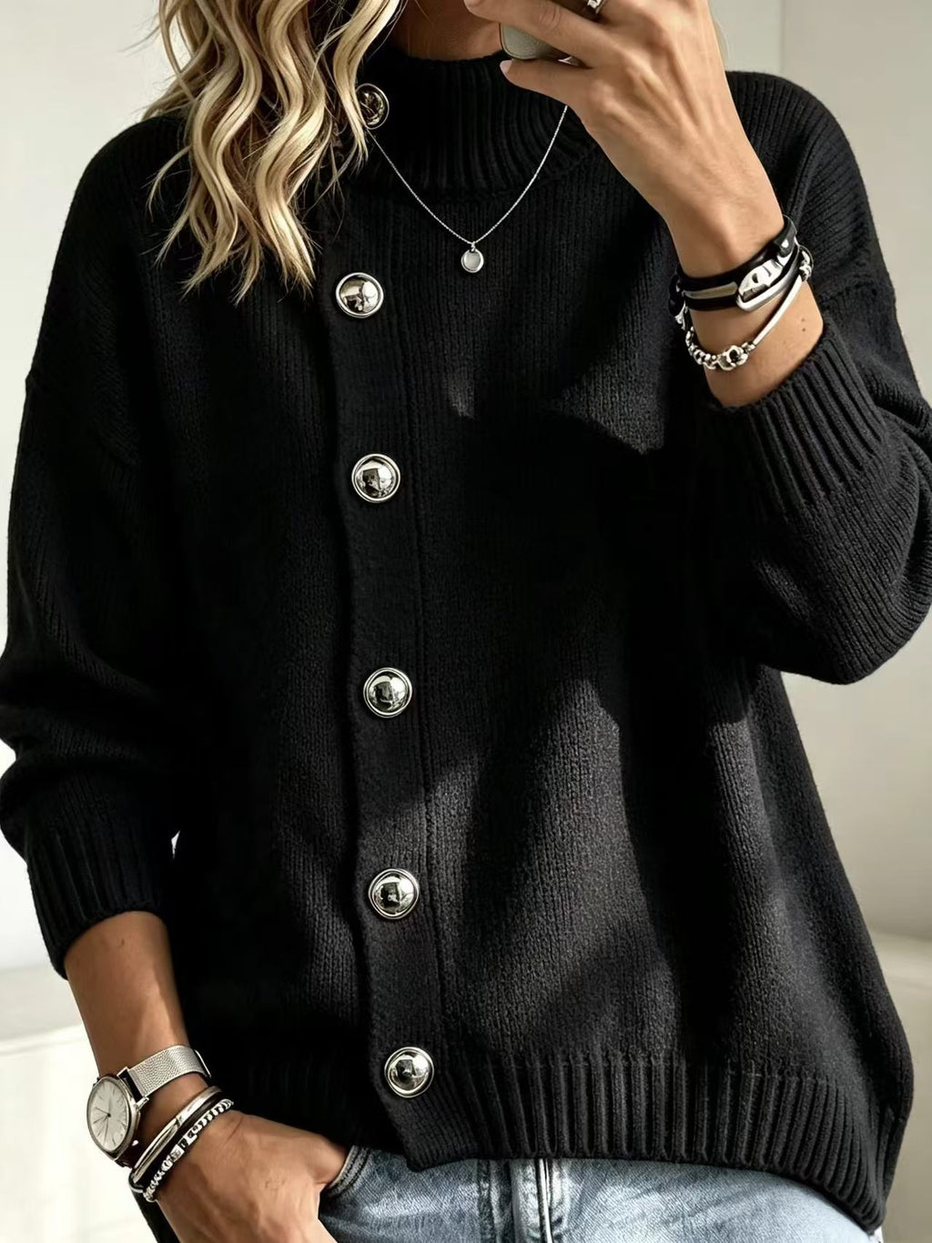 A woman wearing a black ribbed knit cardigan sweater with large silver buttons, paired with light blue jeans and accessorized with bracelets and a necklace.