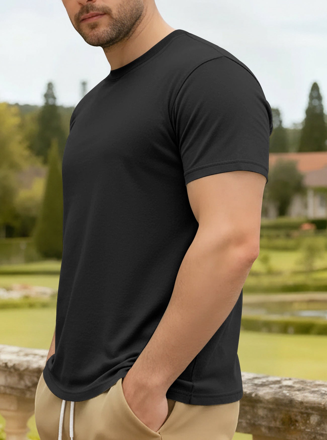 Person wearing a black t-shirt and beige pants standing outdoors with greenery in the background