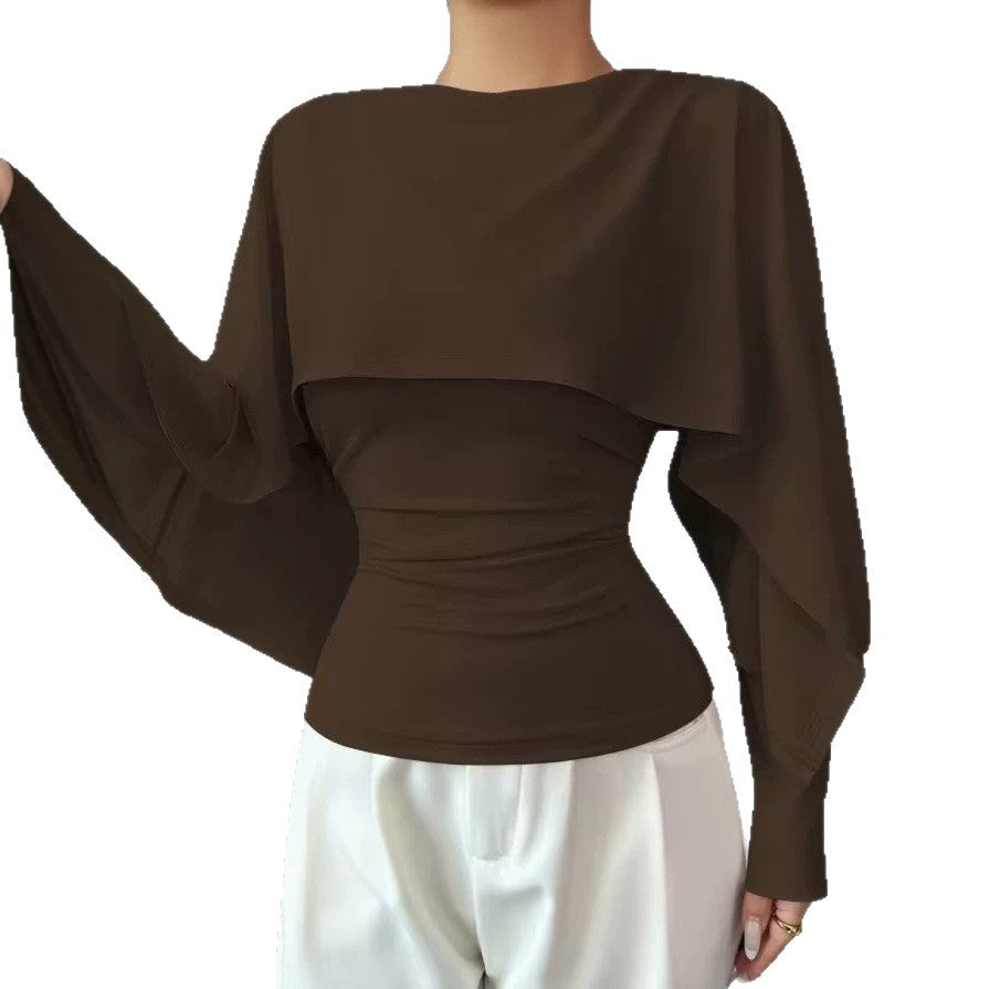 A woman wearing a dark brown long-sleeve top with a draped cape-style collar and gathered waist, paired with white pants, shown against a plain white background.