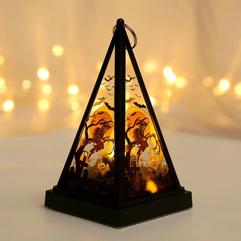 A black pyramid-shaped Halloween lantern with a glowing internal light, featuring cutout silhouettes of a witch on a tree, bats, pumpkins, and a haunted house. The lantern is photographed on a light surface with a warm, blurred bokeh background of string lights.