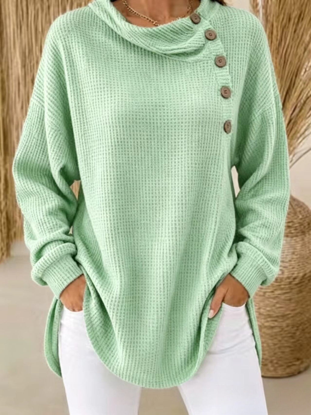 A woman wearing a light mint green waffle-knit cowl-neck sweater with brown buttons down the side, paired with white pants.