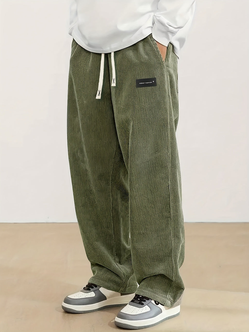 Person wearing green corduroy pants with a white shirt and gray sneakers on a beige background