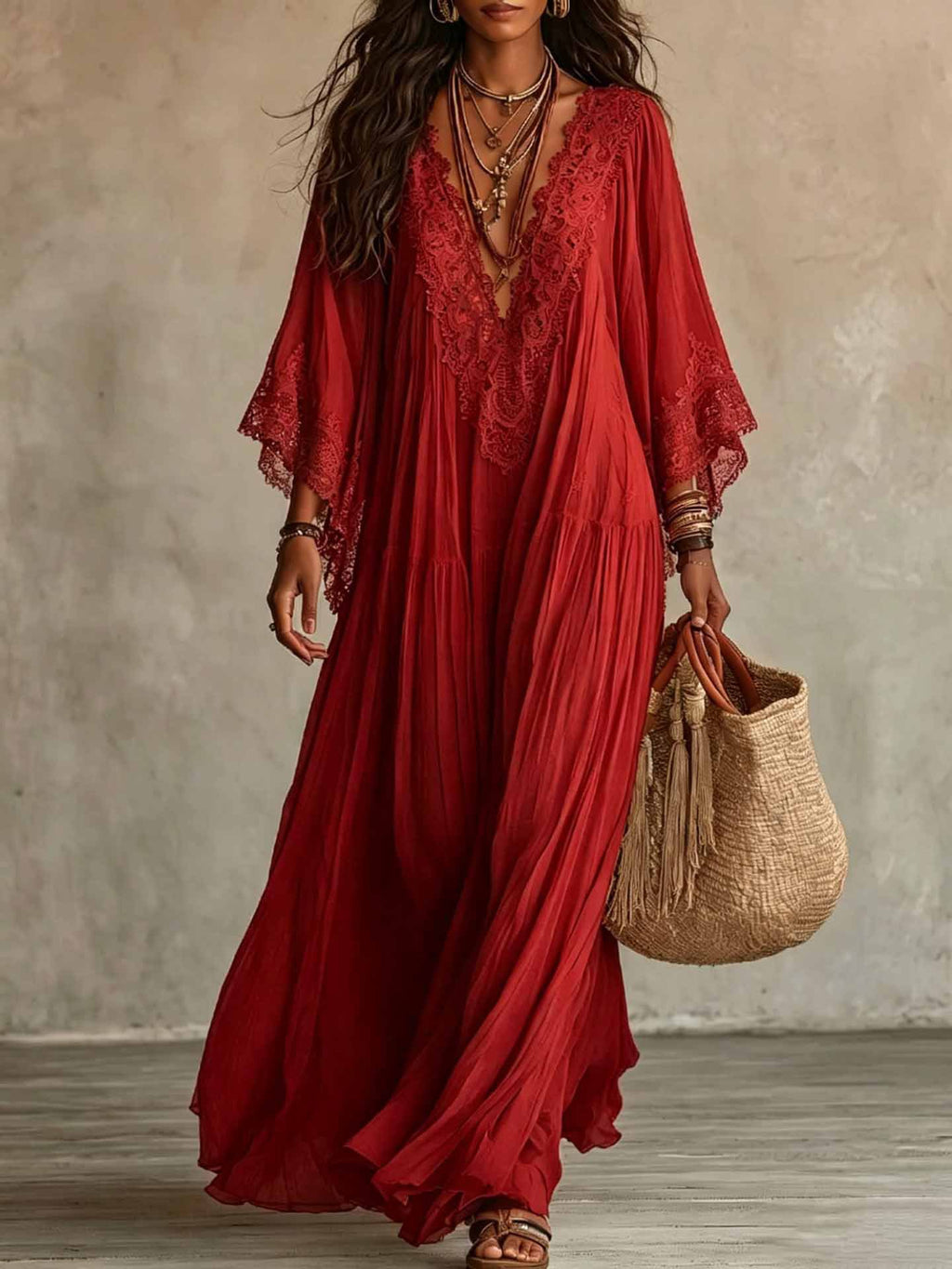 A woman wearing a flowing red maxi dress with lace trim and wide sleeves, holding a woven straw tote bag against a textured neutral wall.
