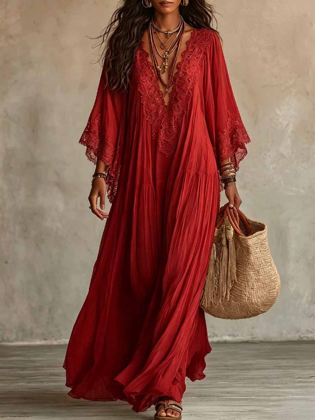 A woman wearing a flowing red maxi dress with lace trim and wide sleeves, holding a woven straw tote bag against a textured neutral wall.
