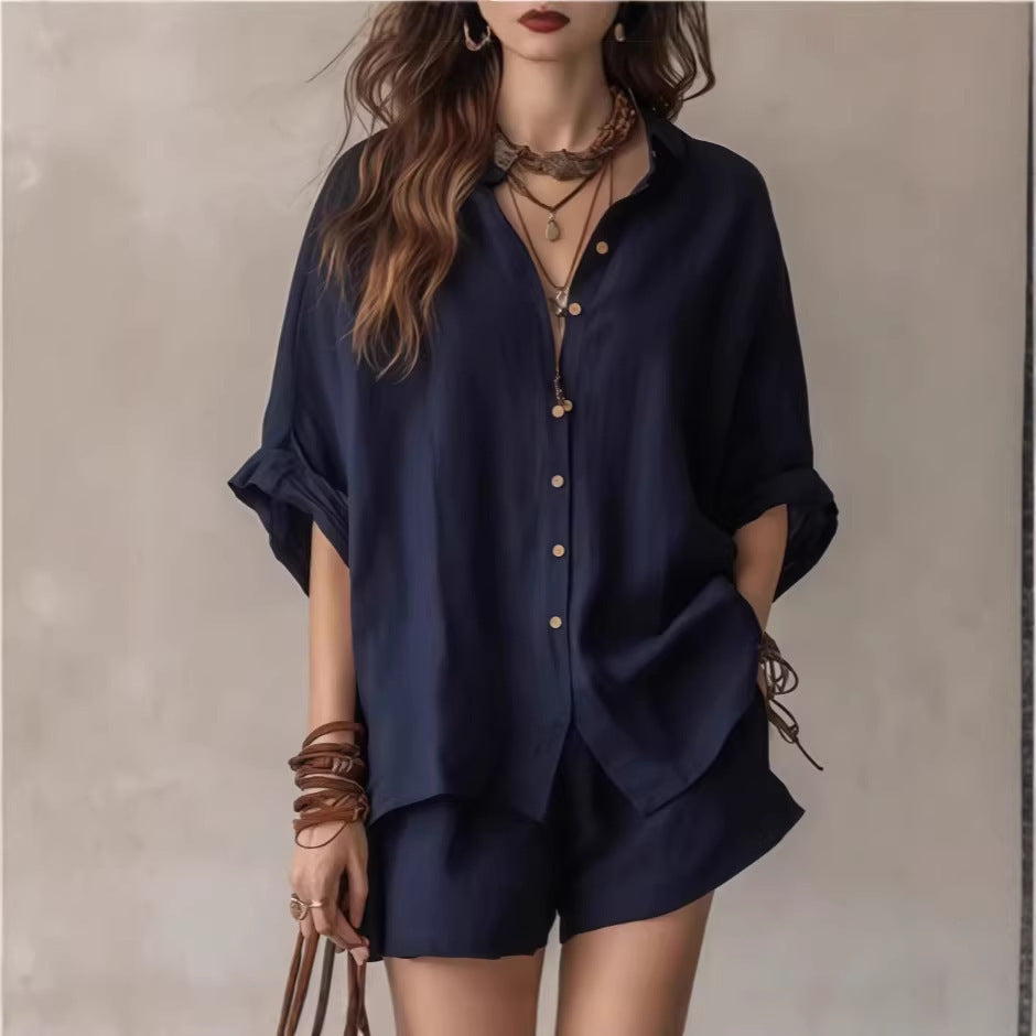 Person wearing a navy blue shirt and shorts with layered necklaces and bracelets on a neutral background