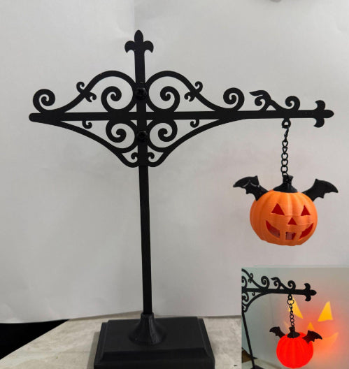 Black metal Halloween display stand with an ornate scrollwork design, holding a small orange jack-o'-lantern pumpkin with black bat wings. A smaller inset image in the lower right shows the stand illuminated in use with a glowing orange pumpkin.