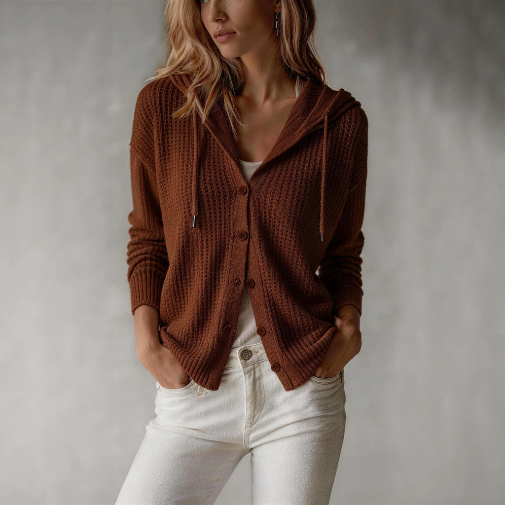 A woman wearing a brown waffle-knit cardigan with a hood and front buttons, paired with white jeans, standing against a textured gray wall.