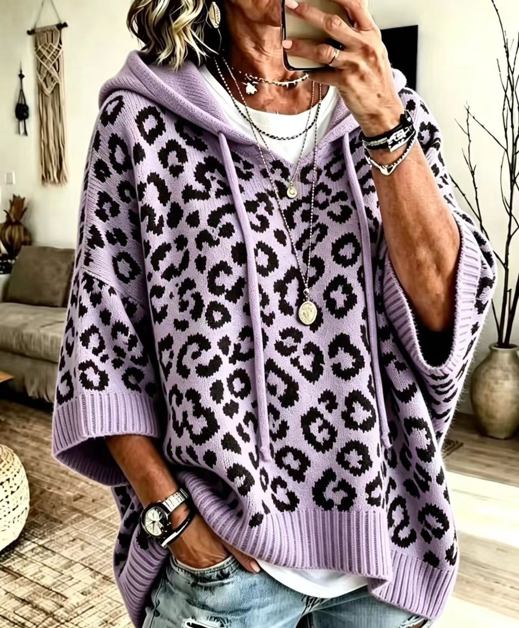 A woman takes a mirror selfie wearing a lavender purple and black leopard print knit hoodie poncho sweater, paired with a white t-shirt and light-wash distressed jeans.