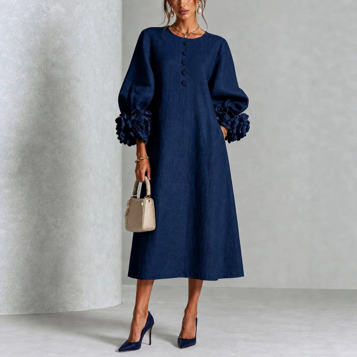 A woman models a dark navy blue midi dress with voluminous long sleeves featuring ruffled cuffs, a round neckline with decorative buttons, and side pockets. She is holding a small beige handbag and wearing navy blue high heels against a textured light gray studio backdrop.