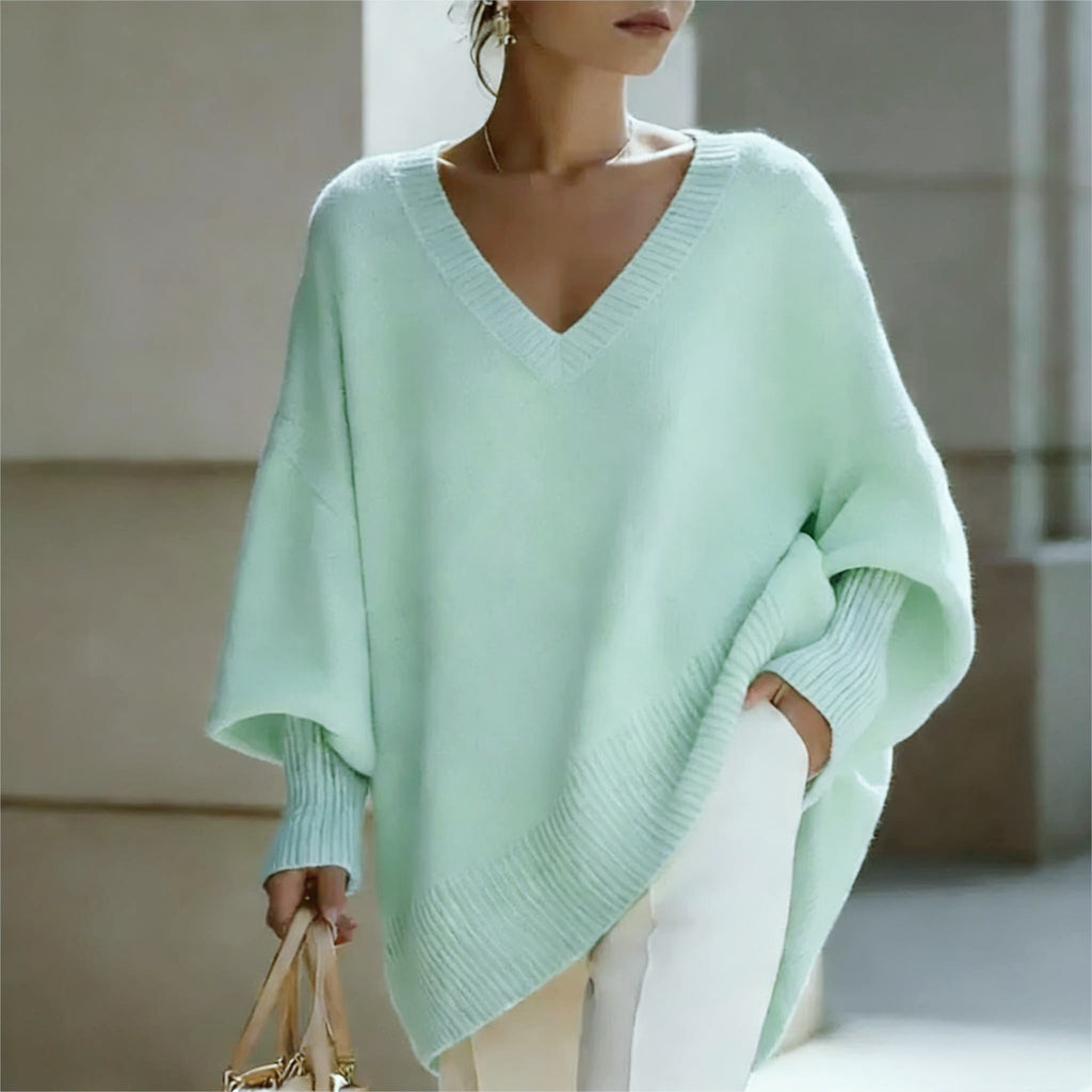 A woman wearing a light mint green knit poncho sweater with a V-neck and wide batwing sleeves, paired with white pants and holding a gold handbag.