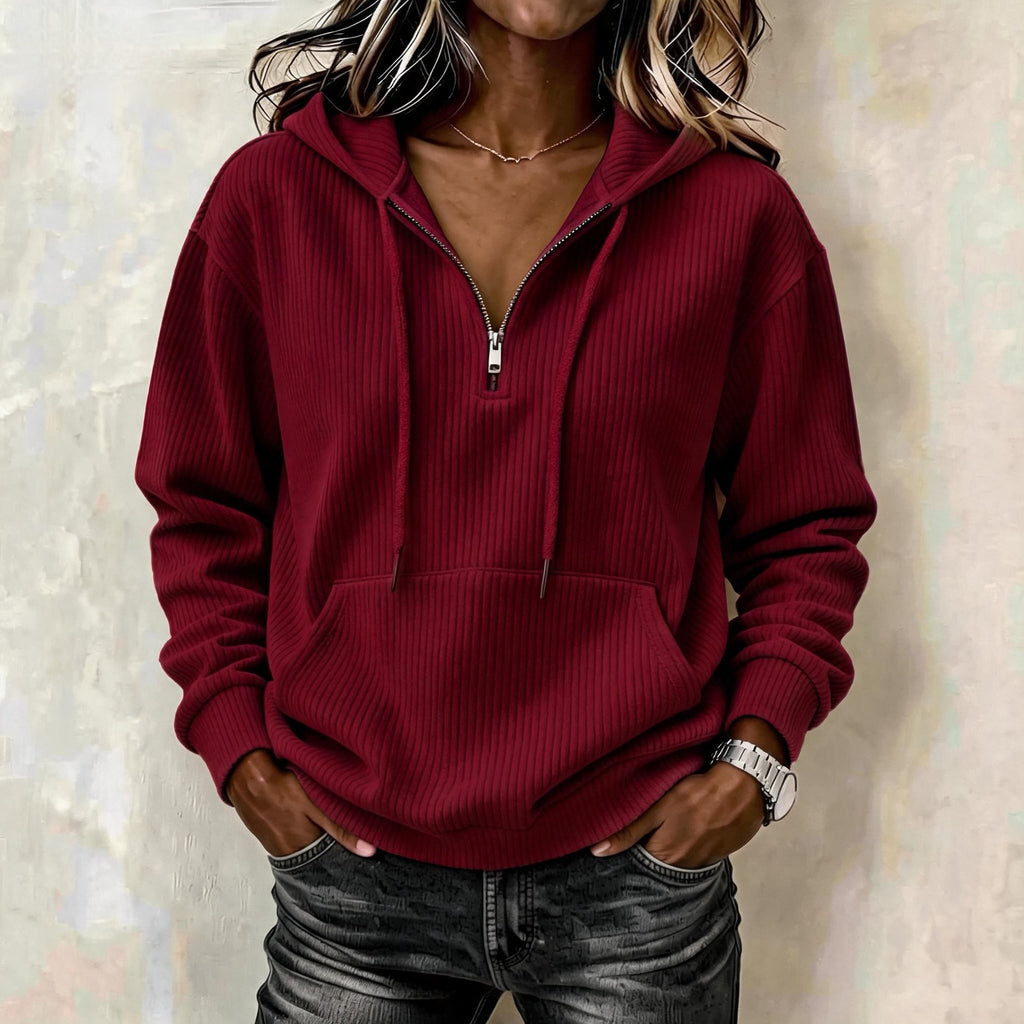 A woman wearing a ribbed half-zip hoodie in deep burgundy red, paired with dark gray jeans, standing against a textured beige wall.