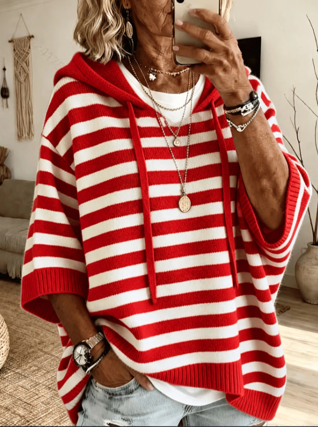 A woman takes a mirror selfie wearing a red and white horizontally striped knit hoodie with wide three-quarter sleeves, light-wash denim jeans, layered gold necklaces, and bracelets in a cozy living room setting.