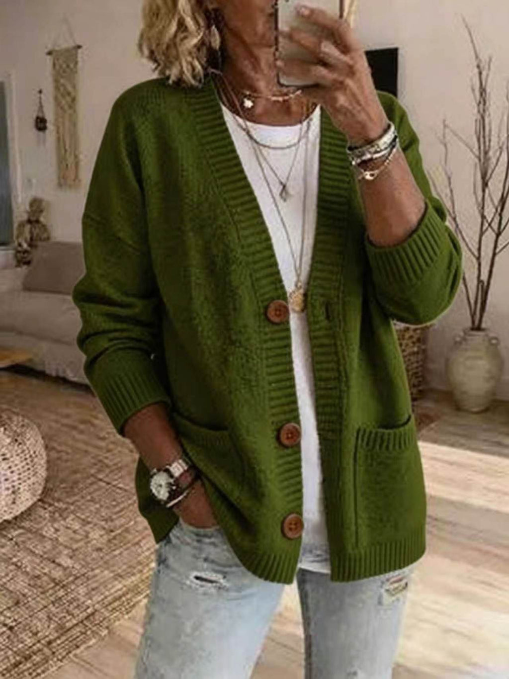 A woman wearing an olive green knit cardigan with brown buttons over a white t-shirt and light-wash distressed jeans, taking a mirror selfie in a cozy living room setting.