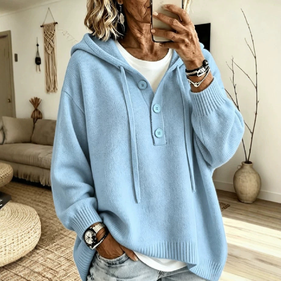 A woman takes a mirror selfie wearing a light blue knit hooded sweater with a buttoned henley-style neckline over a white undershirt, in a cozy living room setting.