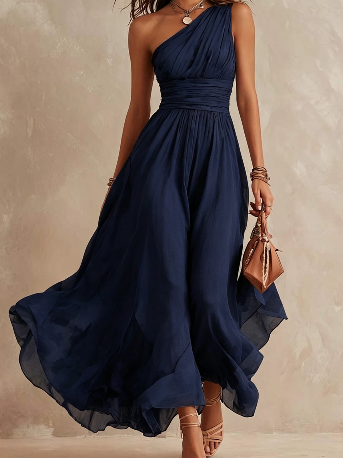A woman wearing a flowing navy blue one-shoulder maxi dress with a gathered waist, holding a small brown handbag against a textured beige studio backdrop.