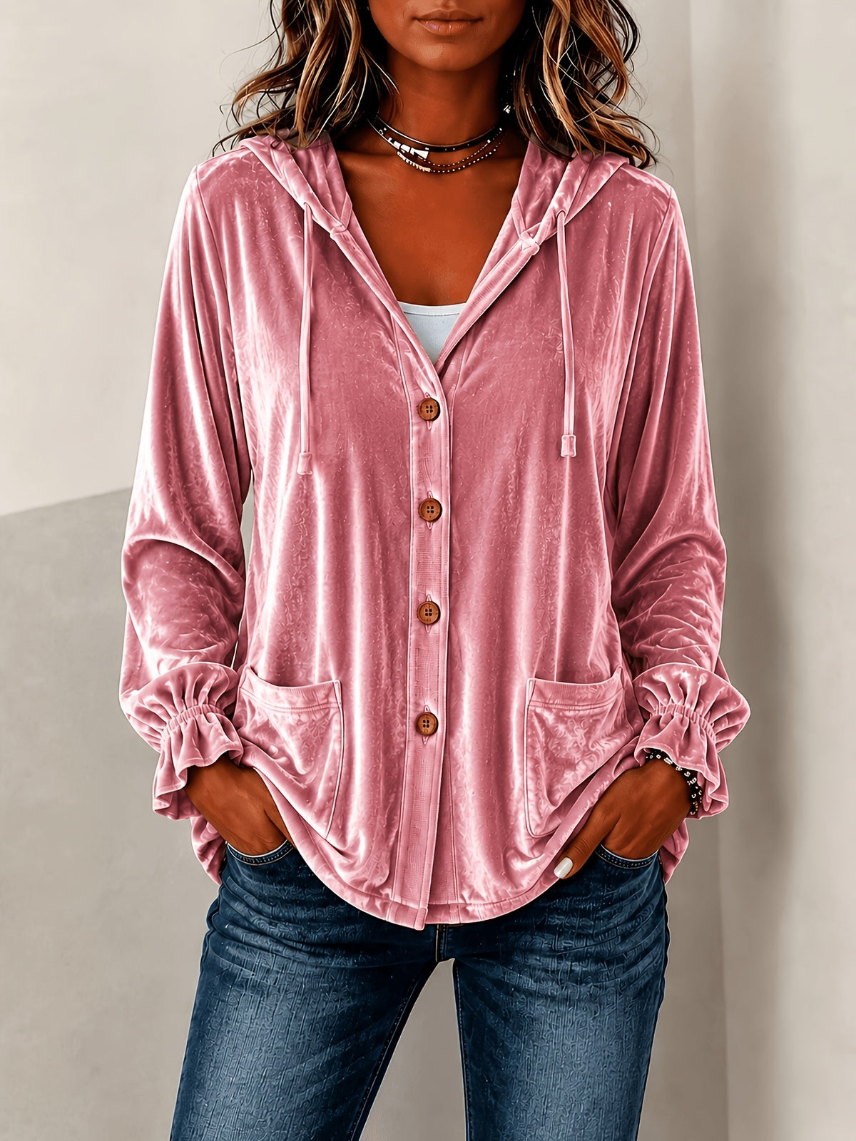 A woman wearing a dusty pink velvet hooded button-up shirt with drawstrings and front pockets, paired with blue jeans.