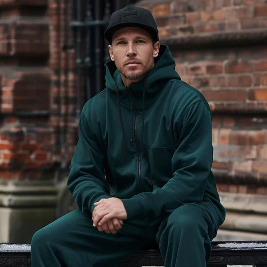 Man wearing a dark green hoodie and pants sitting against a brick wall.